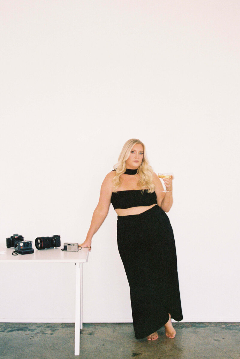 A wedding photographer in a black dress leans on a table covered in cameras while drinking wine