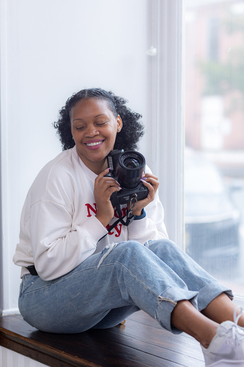 Black women photographer in the window holding a canon r5 camera smiling with her eyes closed. She is a photography studio.
