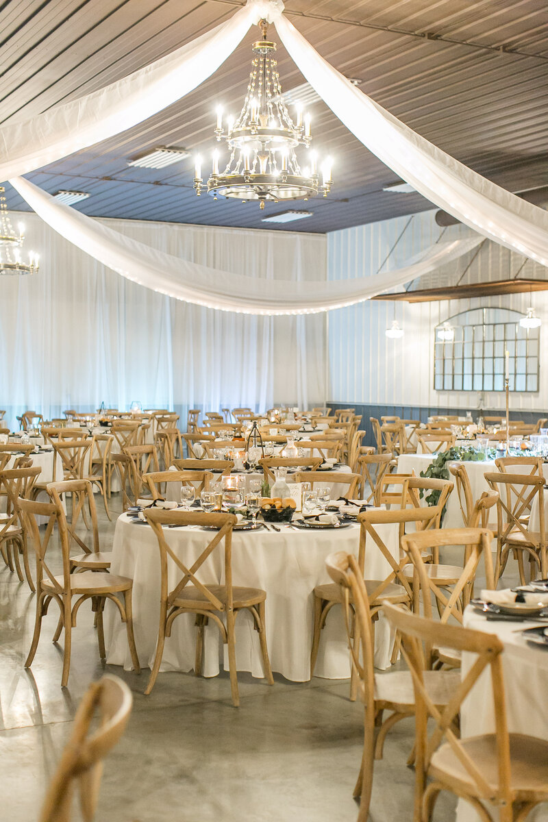 A wedding venue with white fabric draping from the ceiling and hundreds of wooden chairs around white tables