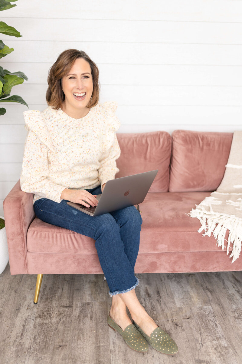 Lynn Davy sitting in pink couch with laptop on lap typing and laughing.