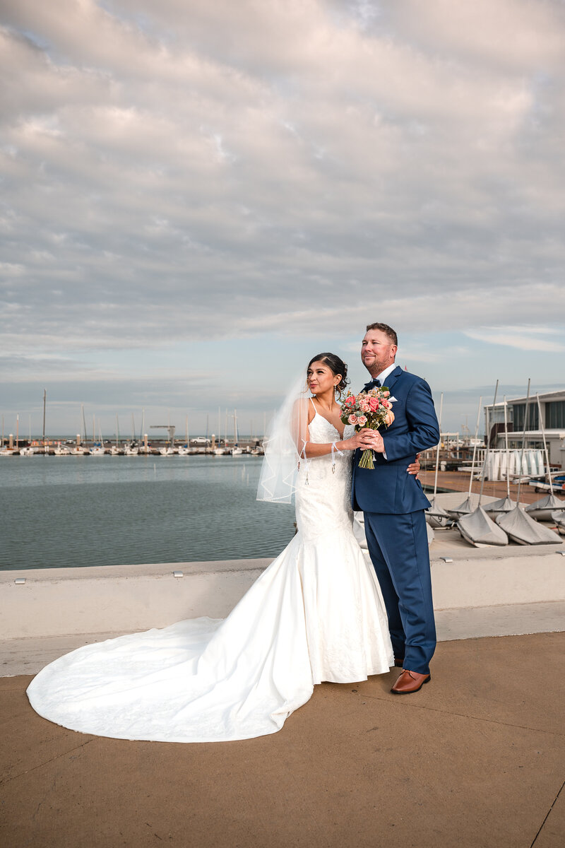 BRIDE AND GROOM STANDING TOGETHER ON THE CORPUS CHRISTI BAYFRONT LOOKING INTO THE DISTANCE WITH PUFFY VIOLET CLOUDS OVER THEM AND BOATS LINED UP BEHIND THEM.