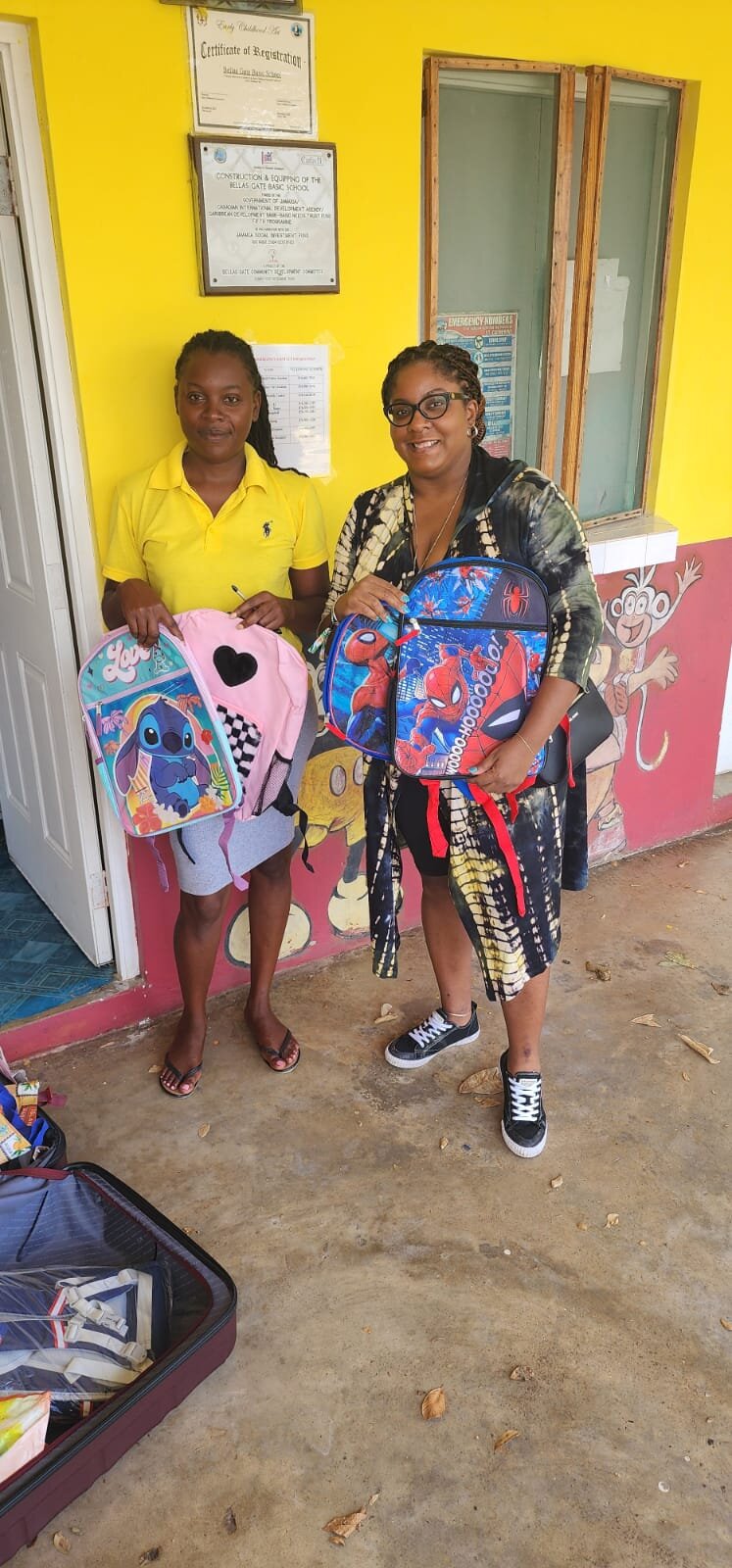 Two women standing in a rural Jamaican school doorway with their arms full of new backpacks from a backpack drive.