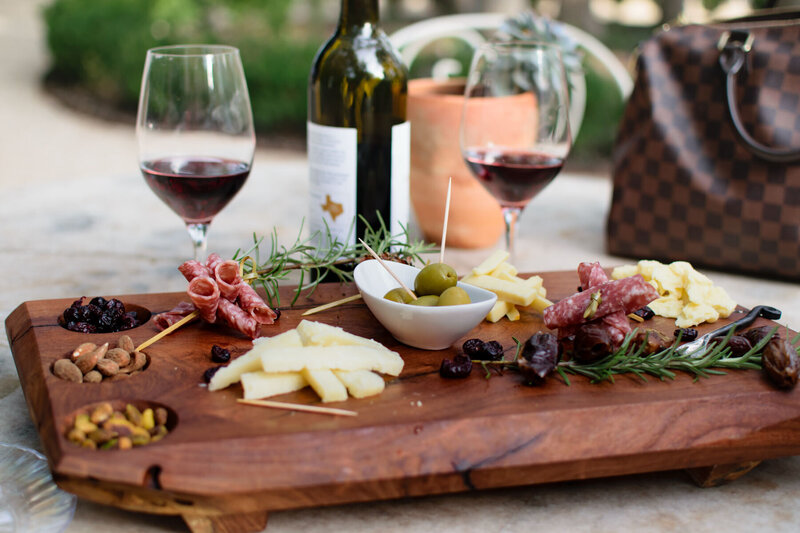 A wooden charcuterie board with sliced cheeses, cured meats, olives, nuts, and dried fruits. Two glasses of red wine and a wine bottle are in the background. A brown checkered bag is placed nearby.