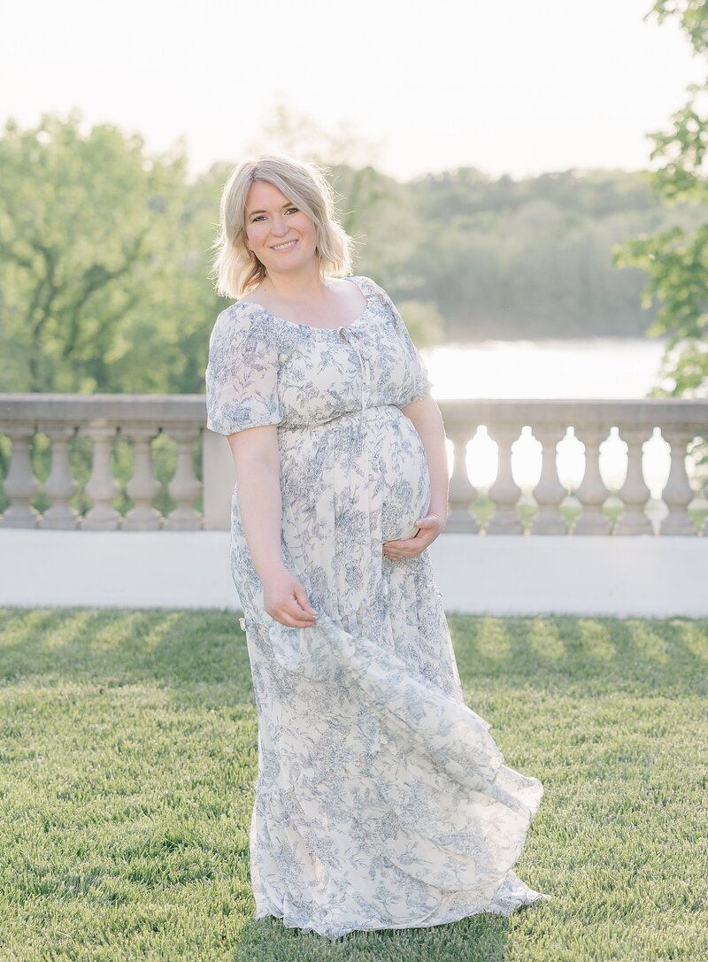 mother in blue floral dress holds baby bump while standing on a grassy terrace overlooking a lake, Indianapolis maternity photographer