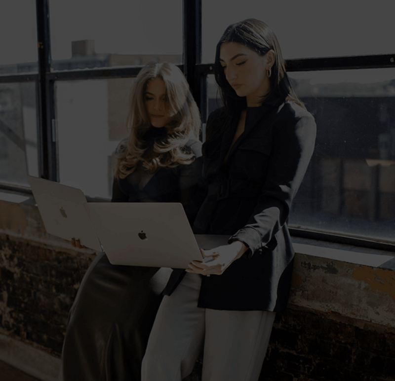 Two women (Isla Luna Co-Founders) leaning against a wall with their laptops working