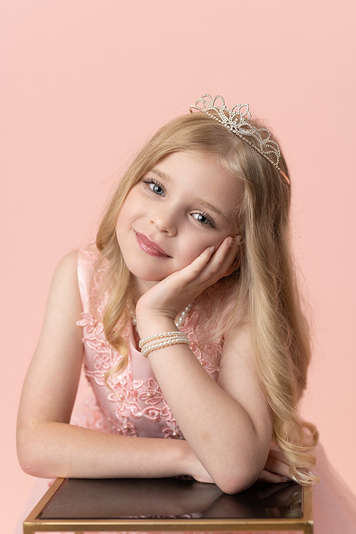 Young girl with long blonde hair, wearing a tiara and lace dress, smiles while holding a bowl of pink macarons. The background is a soft pink.