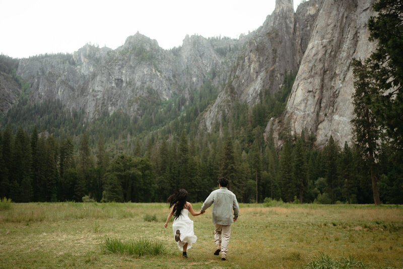 Couple holds hands running into a green meadow surrounded by cliffs, the perfect place for a yosemite wedding