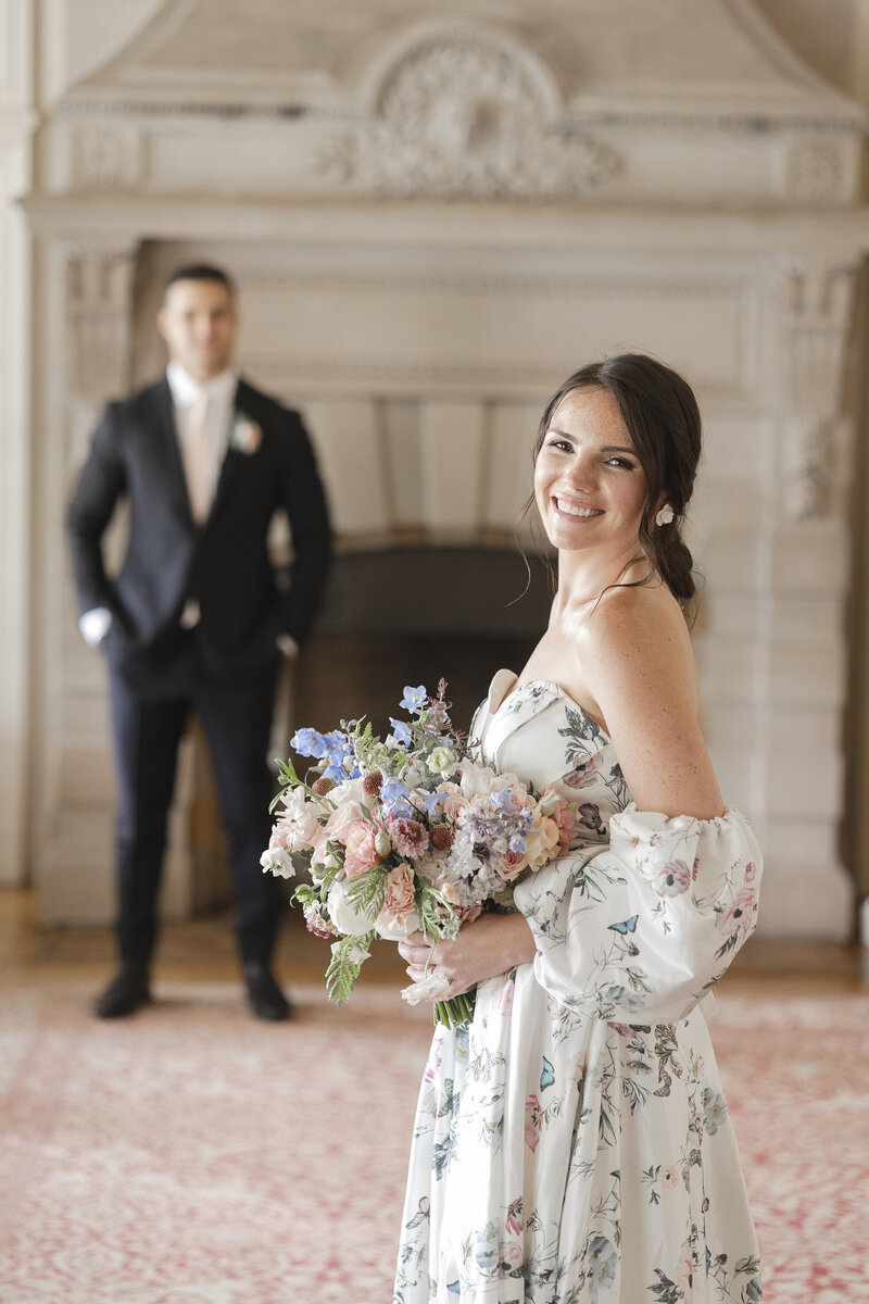 Bride in the foreground smiling at camera, groom in the background looking blurry.