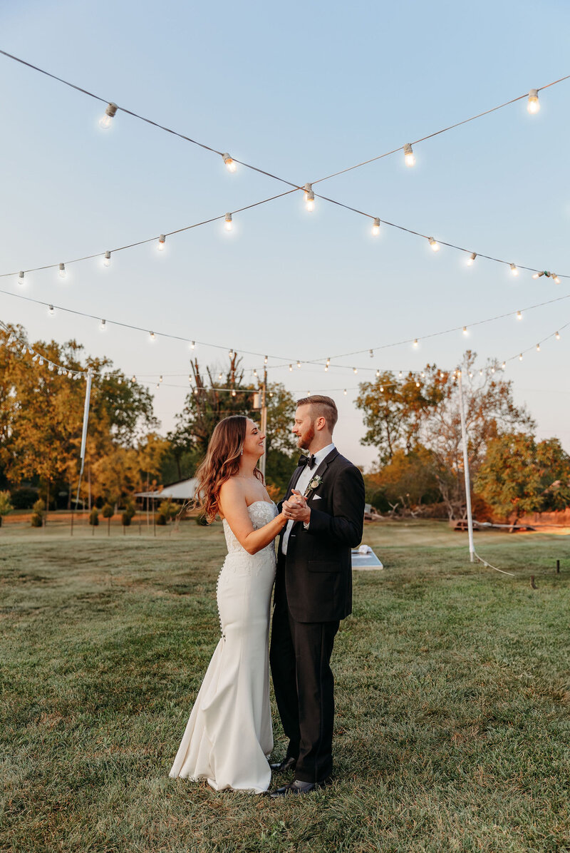 A bride and groom smile at each other during their first dance under string lights at a rustic wedding venue. The bride wears a strapless white gown, and the groom is in a black suit. They are on a grassy field with trees and a clear sky in the background, capturing the essence of an outdoor wedding venue in Virginia.