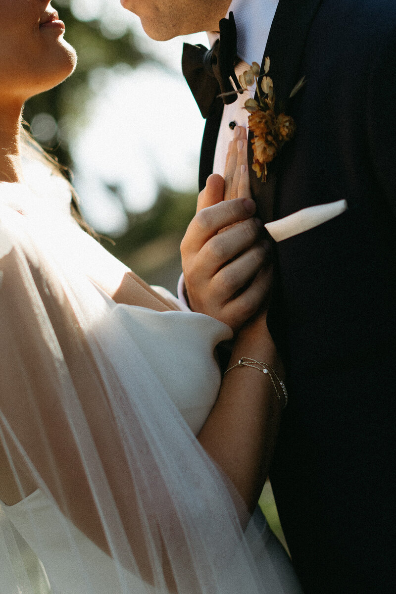 A close-up of a bride and groom facing each other outdoors at their Broadview Hotel wedding, with the bride gently touching the groom's chest near his boutonniere. Both wear formal wedding attire under the warm Ontario sun, beautifully captured by Toronto wedding photographer, Jennifer van Son.