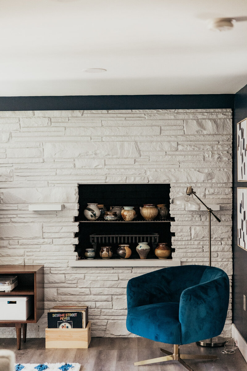 A living room featuring a bold blue velvet chair, a mid-century modern side table, a record crate, and decorative pottery displayed in a white-painted brick fireplace.