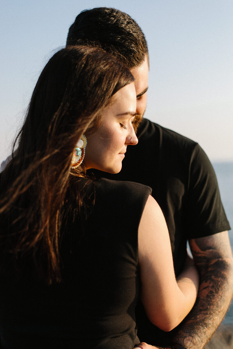 A woman and man embrace closely by the seaside, captured beautifully by a Toronto engagement photographer, Jennifer van Son. She has long brown hair and a colorful earring, and both are wearing black shirts.