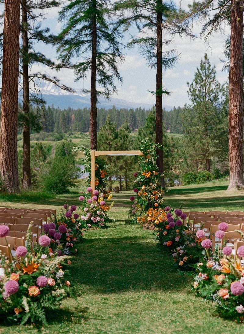 Forest wedding ceremony aisle lined with vibrant garden-style florals in purple, orange, and pink leading to a modern wooden chuppah, designed by Summer Robbins Flowers in Central Oregon