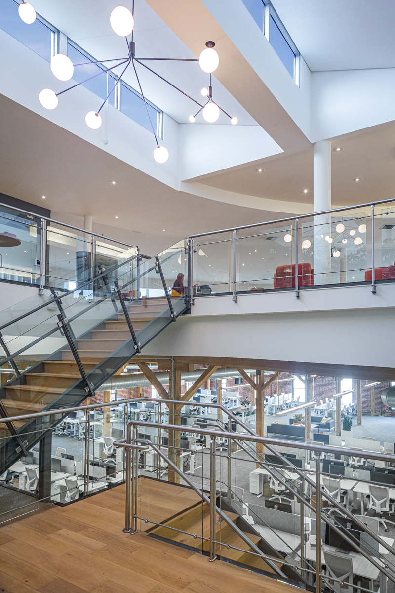 Open-concept workspace with exposed wood beams, large staircase, and sleek glass railings connecting two office levels.