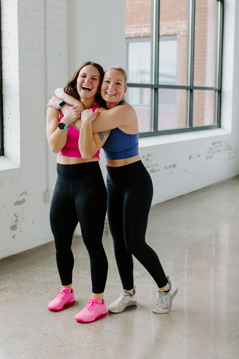 2 women, 1 blonde, 1 dark hair in black leeggins and sports bras hugging