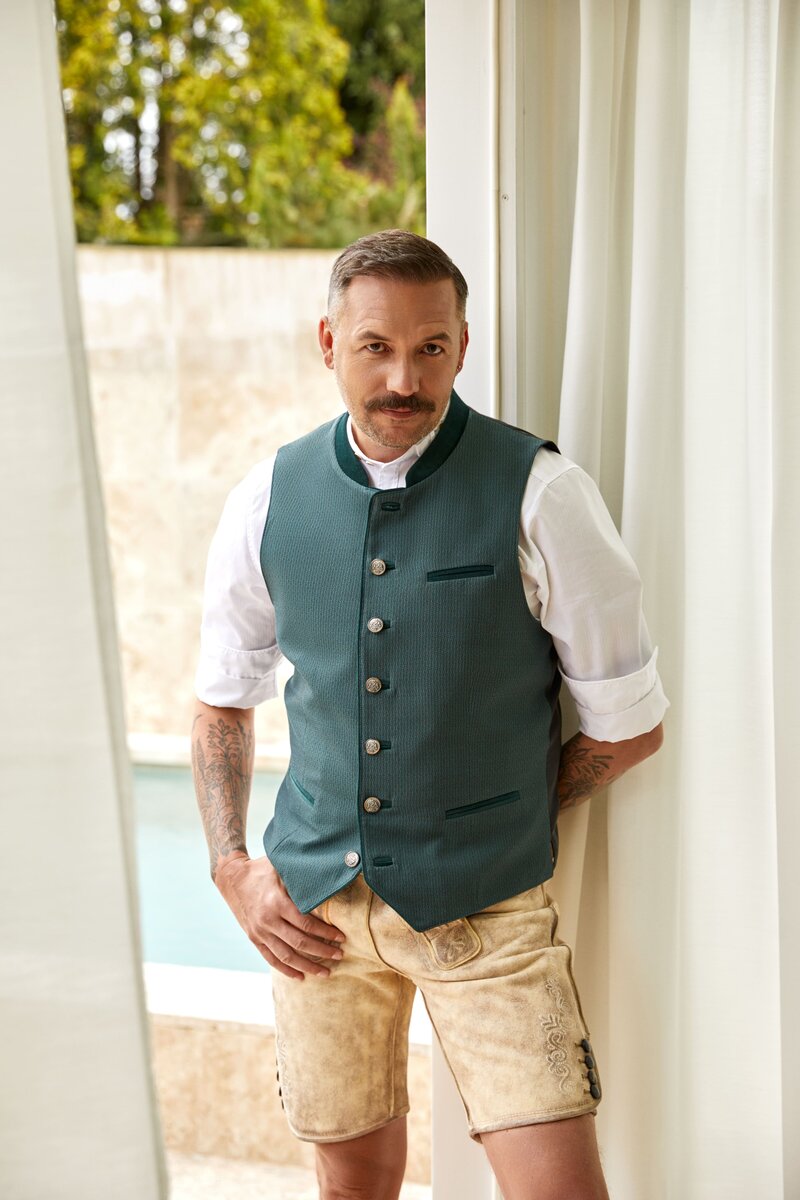 Mann in grüner Designer-Trachtenweste von Monaco Spatzl, kombiniert mit traditioneller kurzer Hirschlederhosen. Die moderne Weste trifft auf klassische Farben und sorgt für einen eleganten, zeitgemäßen Look. Bayerische Tradition modern interpretiert vom Designlabel Monaco Spatzl. 
