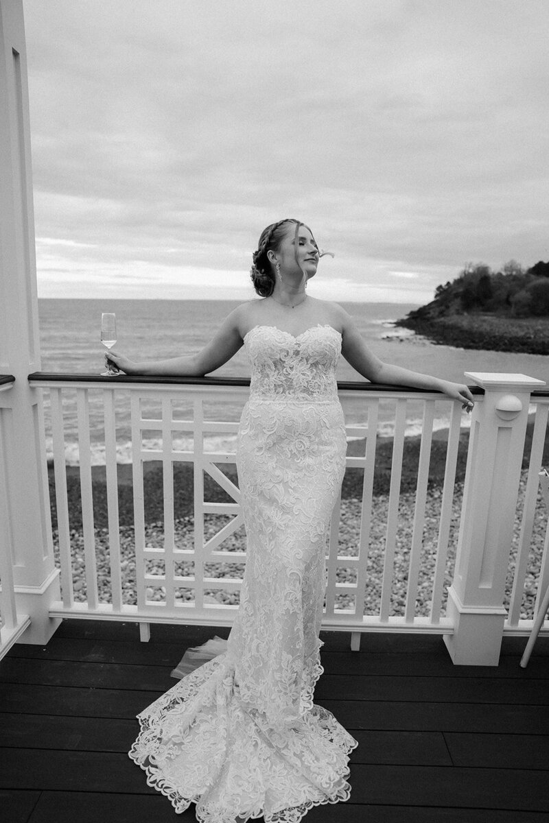Bride leans on the railing overlooking the Atlantic along the MA coastline in her dress with champagne in hand. 