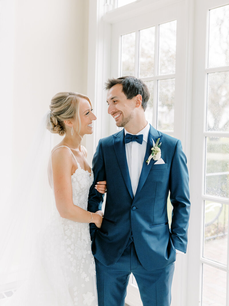 Bride and groom standing together smiling at each other beside a window