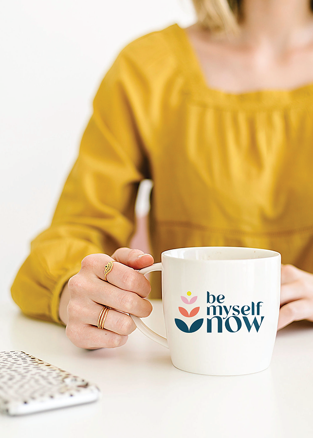 Woman in yellow shirt holding white mug, featuring the Be Myself Now logo, resting on white desktop