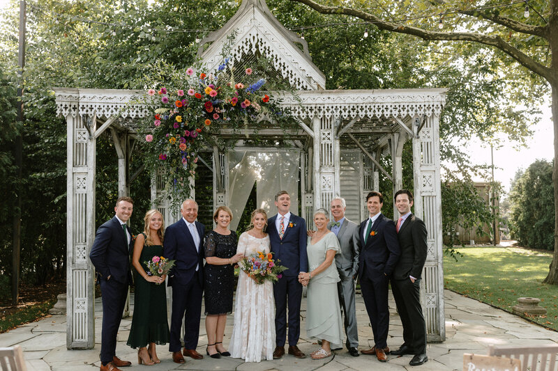 a family  poses under a floral arch at an outdoor venue, featuring a charming structure surrounded by greenery.