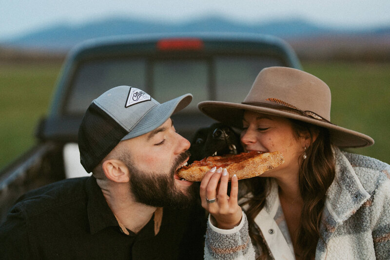 Couple and their black dog sitting in the bed of an antique truck, all biting the same slice of pizza.