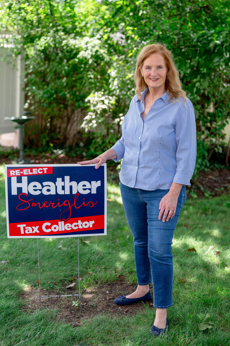 Blonde woman standing outside, smiling at the camera, with her hand on a candidate sign saying "Re-elect Heather Smeriglio - Tax Collector"