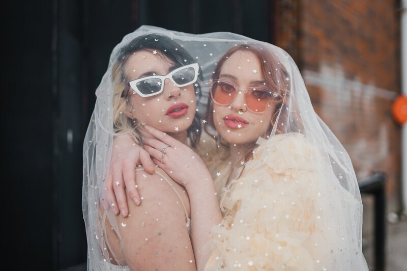 female bridal couple embracing under a veil  looking at the camera.