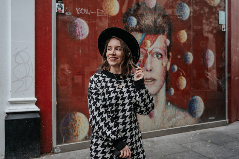 A woman wearing a check jumper and black hat infront of Bowie mural