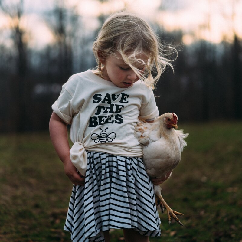 A small blonde girl is wearing a 'Save the Bees' t-shirt and holding a chicken.
