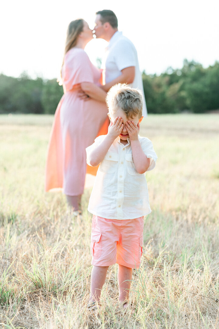 a toddler plays peek-a-boo, covering his eyes while his pregnant mother and his father kiss in the background at Brushy Creek Lake Park. 