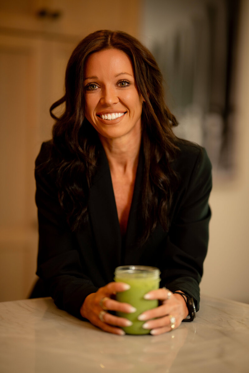 Kenzie Mosher smiling in a black blazer while holding a green smoothie at her kitchen counter — Functional Diagnostic Nutrition Practitioner and women’s health expert.