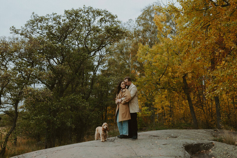 Couple standing and hugging their dog standing besides them trees in fall colours behind the in Lauttasaari in Helsinki