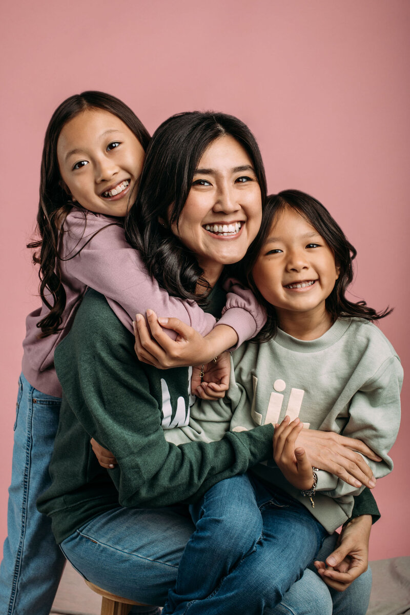 Mother poses with her two daughters in front of a pink background for a family photo session in Emeryville, CA