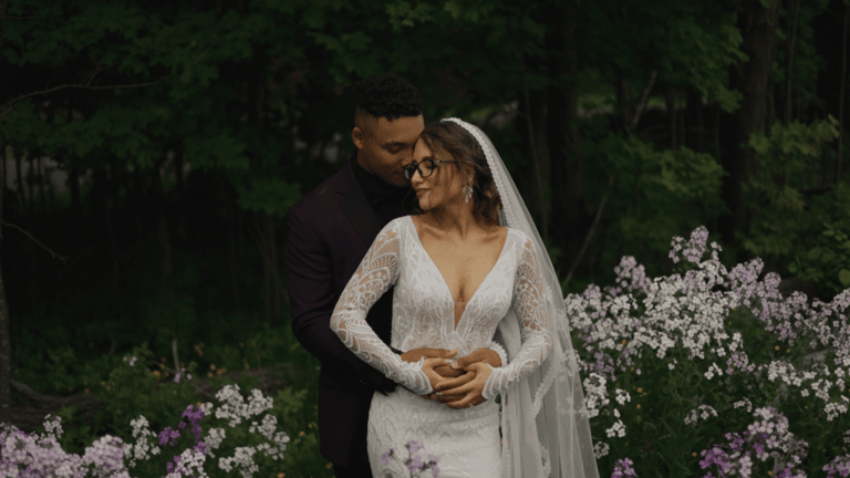 Newlywed couple, groom in a purple suit and bride in a long sleeve, lace, detailed dress, with a long veil, standing together with their heads touching in a small field of purple and white flowers in front of trees