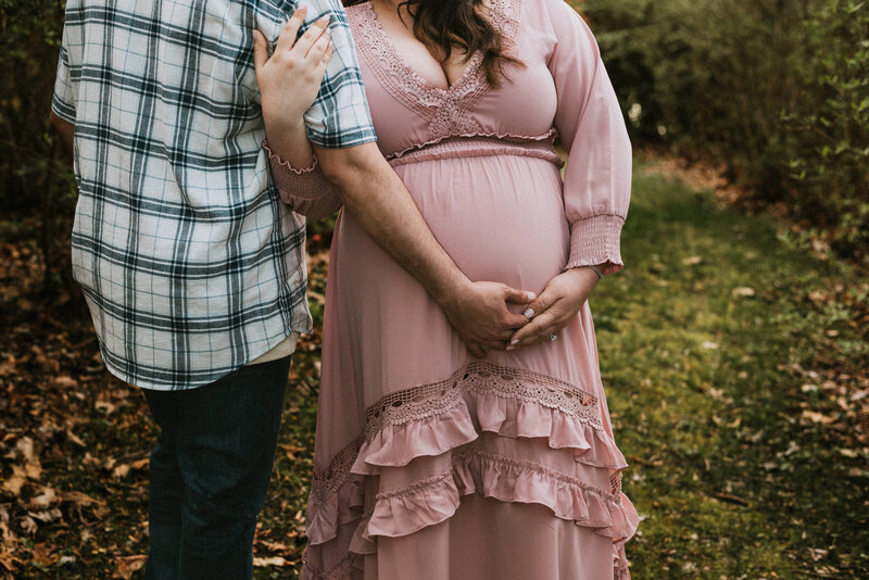 Motherhood photo of belly picture of mom in pink dress holding dads arm and her belly facing the camera while dad is in blue and white plaid facing away from camera holding moms belly in Maryland.