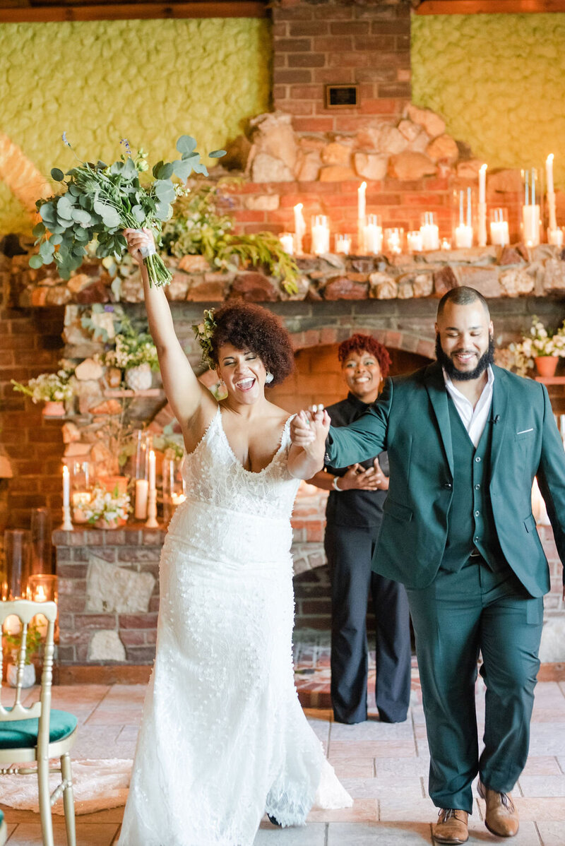 Newlywed couple walks hand-in-hand, smiling joyfully in front of a candlelit stone fireplace in the century room at grbic events as the bride raises her bouquet in celebration.