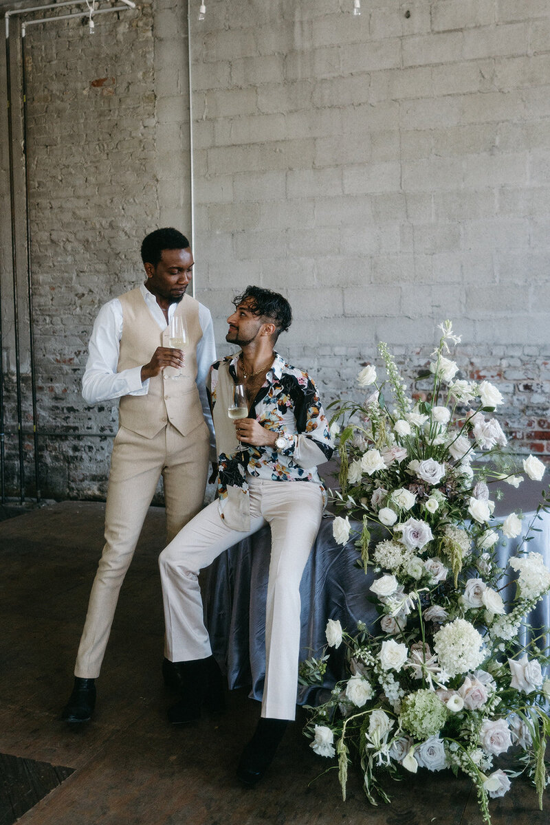 Two men in stylish outfits with champagne, floral arrangement.