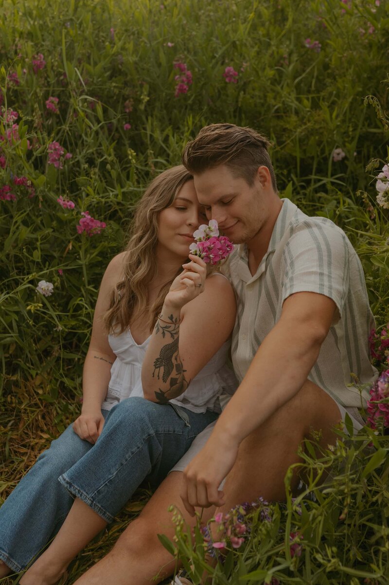 A couple sits closely together in a lush meadow filled with wildflowers, sharing a tender moment. The woman gently holds a small bouquet of pink blooms near her face, while the man leans in with a soft smile. Their casual attire and relaxed pose highlight their connection amidst the natural beauty.