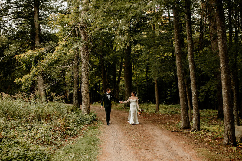A bride and groom stroll down a dirt path below towering pines following their lakeside wedding ceremony.