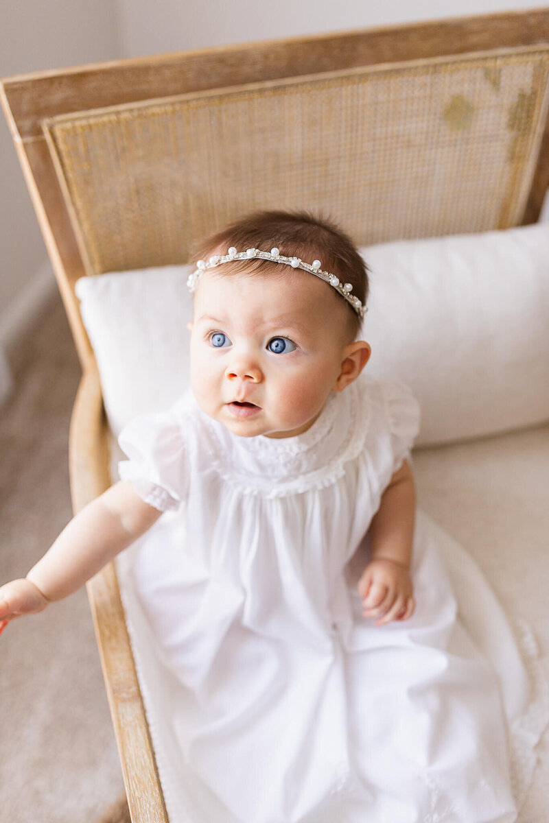 6 month old baby girl with bright blue eyes in a white baptismal dress and a pearl headband