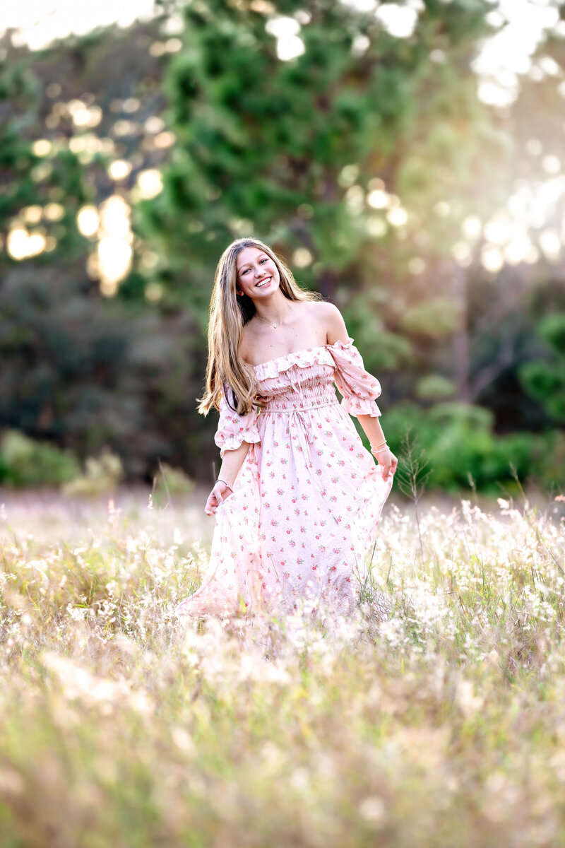 High school senior in a pink floral dress smiling in a golden field during a spring portrait session in Winter Garden, Florida.
