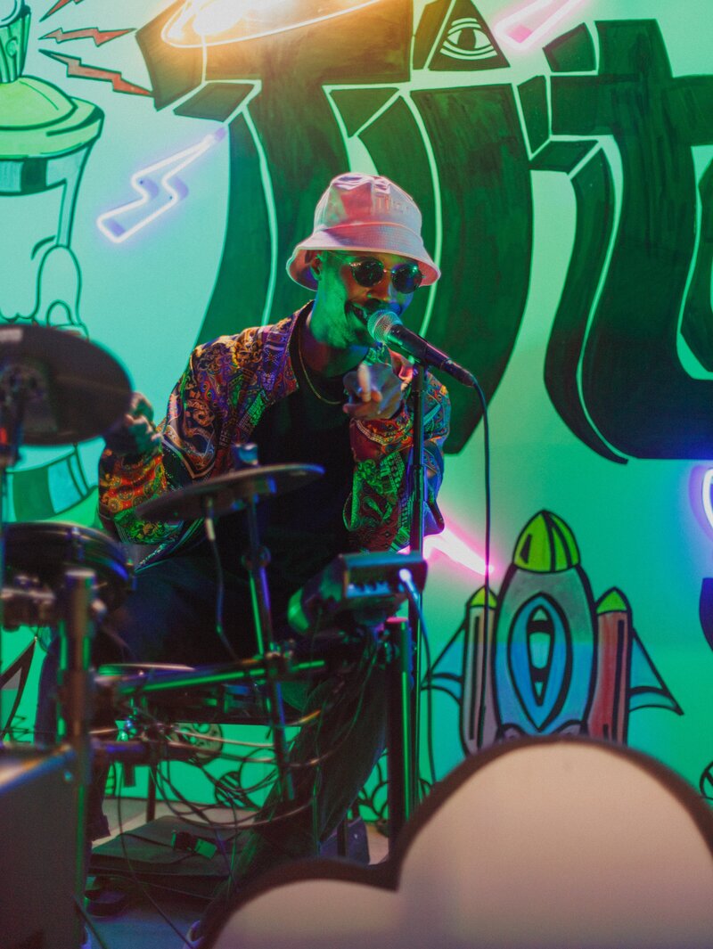 Music performer wearing sunglasses, a pink bucket hat, and a colorful patterned jacket sings into a microphone during Titan Day 3 in New York, with neon lighting and bold graffiti-style wall art in the background.