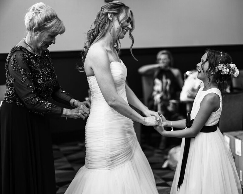 Bride holds hands with flower girl as her mother buttons up  bride's dress