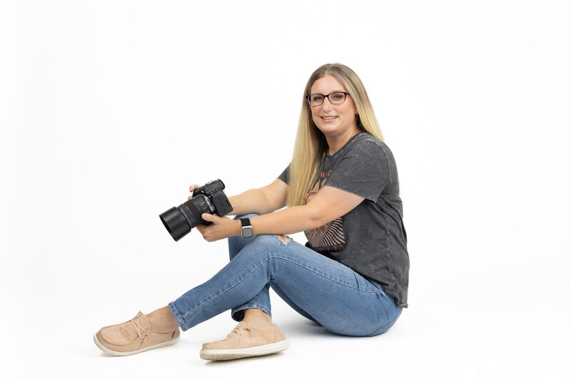 Megan Bowers, a professional senior photographer, poses confidently with her camera against a clean white backdrop. Her relaxed and approachable style reflects her passion for capturing authentic and expressive moments. Megan Bowers Photography specializes in bold and timeless senior portraits.