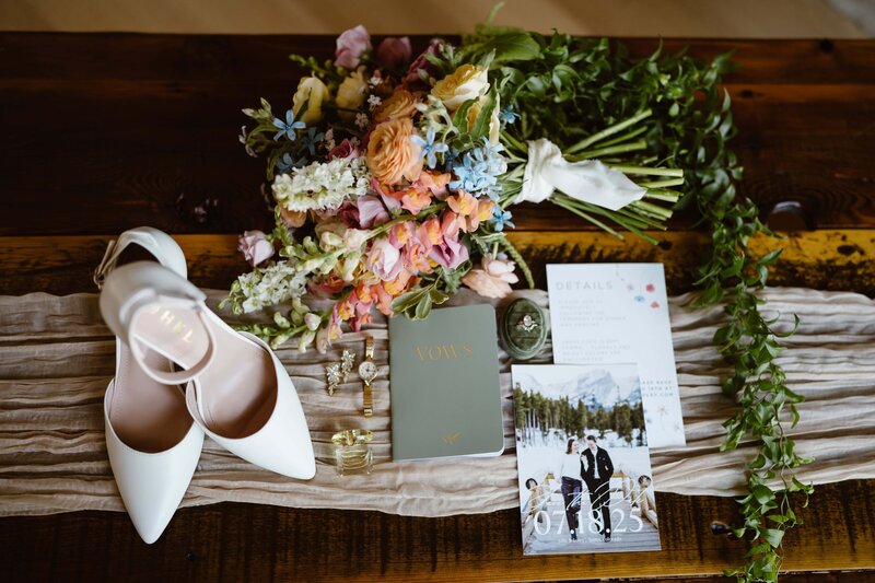 Detail photo of brides white high heels, colorful bouquet, vow book, jewelry, and invitations taken by Colorado wedding photographer Avenir Photo Co.