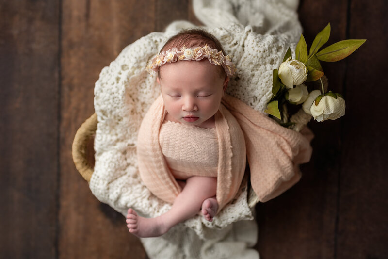 Newborn girl posed in a peach wrap, lying among lace textures and surrounded by delicate florals.