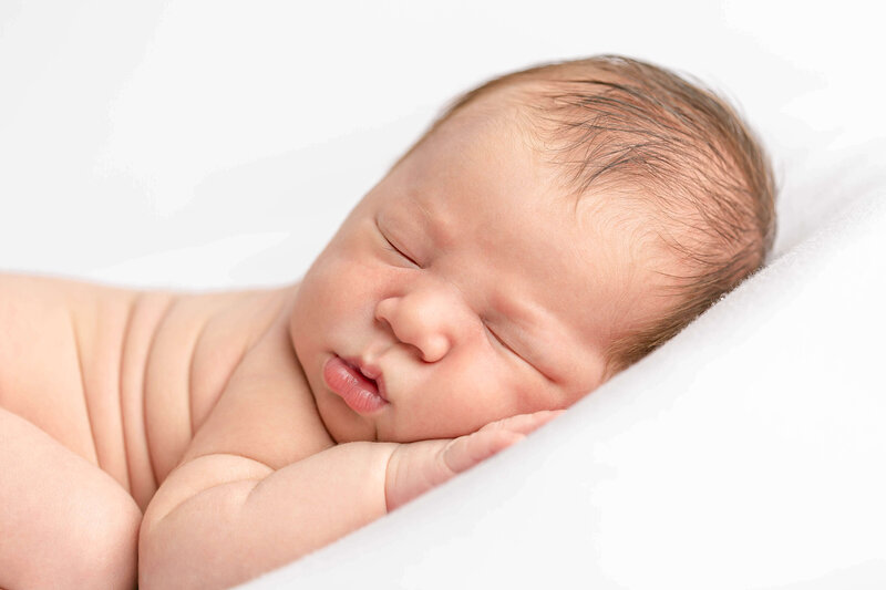 Newborn baby sleeping peacefully on stomach with hand up by face and laying on a white blanket.