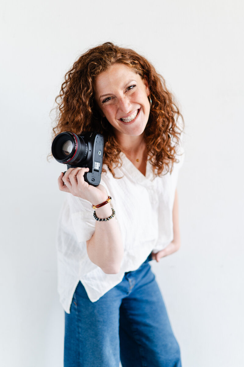 Headshot of Jackie Schon Pierce from LightPop Studio holding a camera against a white backdrop.
