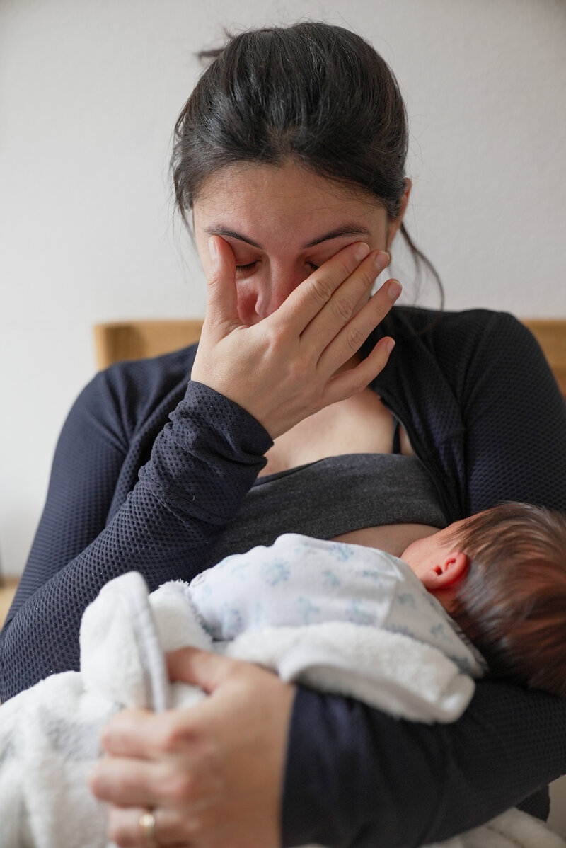 Mother breastfeeding her newborn with a hand on her face, appearing tired or emotional, representing the challenges and support needed during the breastfeeding journey.