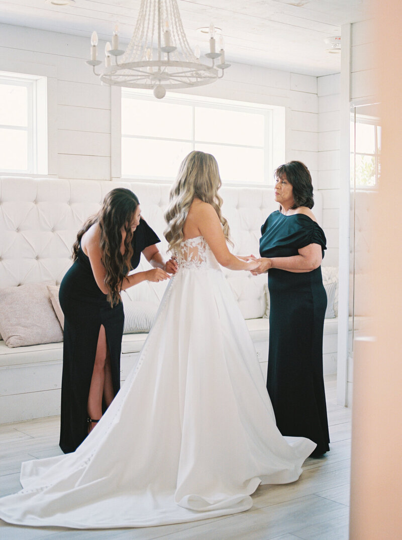 Mother of the bride and maid of honor help the bride get dressed into her wedding gown